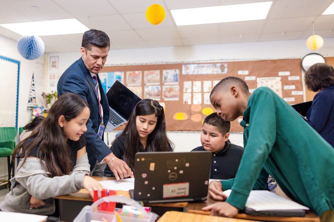 An educator working with a small group of elementary students on laptops